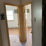 View of an open door leading to a hallway with two adjacent doors, both open, and floors partially covered with paper and blue tape. Natural light comes in through a window.
