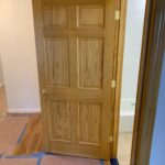 A wooden interior door is partially open between a hallway with paper-covered floors and a bathroom with a white bathtub.