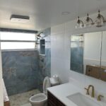 Modern bathroom with marble-tiled shower, glass door, gold fixtures, toilet, dark wood vanity, white countertop, and large mirror above the sink.