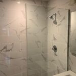Bathroom with white marble-patterned tiles, a glass shower partition, a wall-mounted showerhead, and a mirror above a sink.