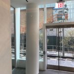 A white cylindrical column stands near the glass entrance doors of a modern building, with large windows showing an outdoor sidewalk and construction barriers.