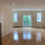 Bright, empty room with light wood floors, beige walls, two windows, and a glass door leading to a small balcony with a view of green trees and a building.