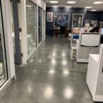 Modern office hallway with glass-walled rooms, desks, computers, printers, and office supplies; tiled ceiling and polished concrete floor.