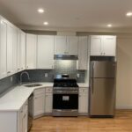 Modern kitchen with white cabinets, stainless steel appliances, gray subway tile backsplash, and wooden floor. Refrigerator, oven, and dishwasher are visible.