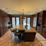 Spacious kitchen with dark wooden cabinets, granite island with chairs, pendant light fixture, and large windows leading outside.