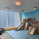 Indoor playroom with padded mats, climbing structures, slide, bean bag chairs, and large windows with city building views.