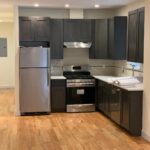 Modern kitchen with dark cabinets, stainless steel fridge and oven, white tile backsplash, and hardwood floors. A sink and countertop are positioned under a small window.