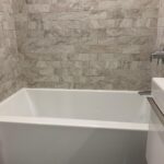 A white bathtub with beige stone tile walls, a silver faucet, and part of a white sink visible on the right.