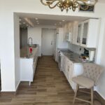 Modern kitchen with white cabinets, marble countertops, and wood-look tile flooring. A beige upholstered chair sits near the counter; chandelier and cleaning supplies are visible.